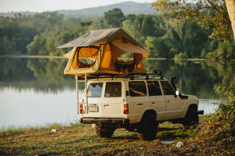 white off road van with an orange tent on the roof, parked next to an open lake