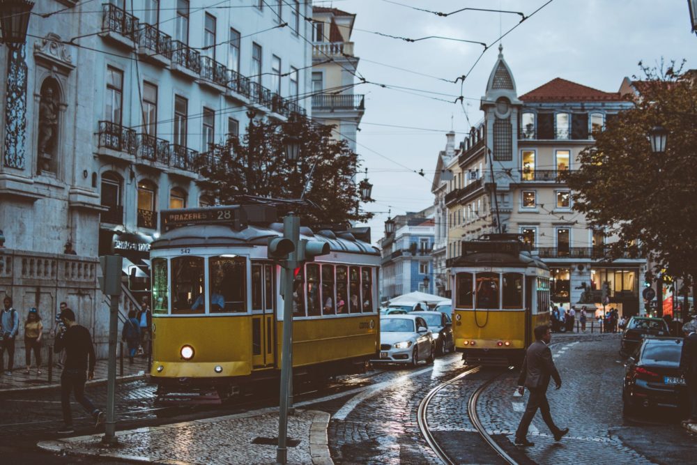 Two bright yellow trams, driving past pedestrians and ornate architecture in Lisbon, Portugal