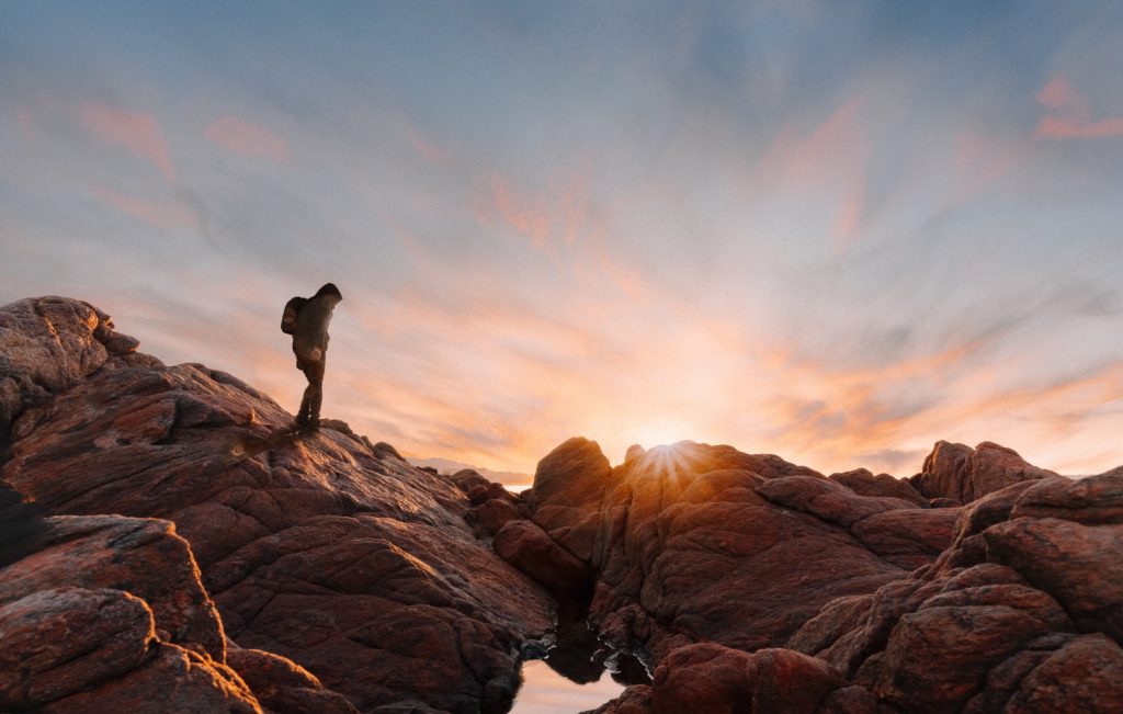 The silhouette of a backpacker climbing over rocks in the pink hues of a sunset.