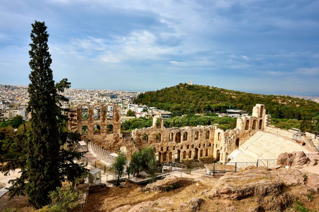 a landscape of Athens with the ruins of an ancient Greek theatre in the foreground and green forests, the city and the sea in the distance.