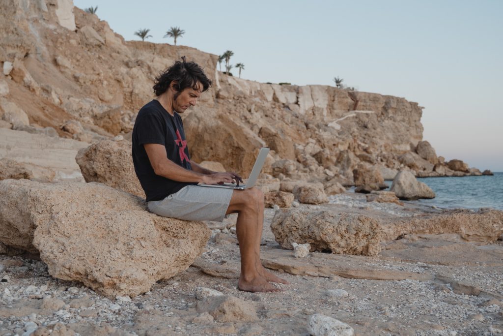 A man sits on a rock at the beach on his laptop, making the most of the workcation travel trends.