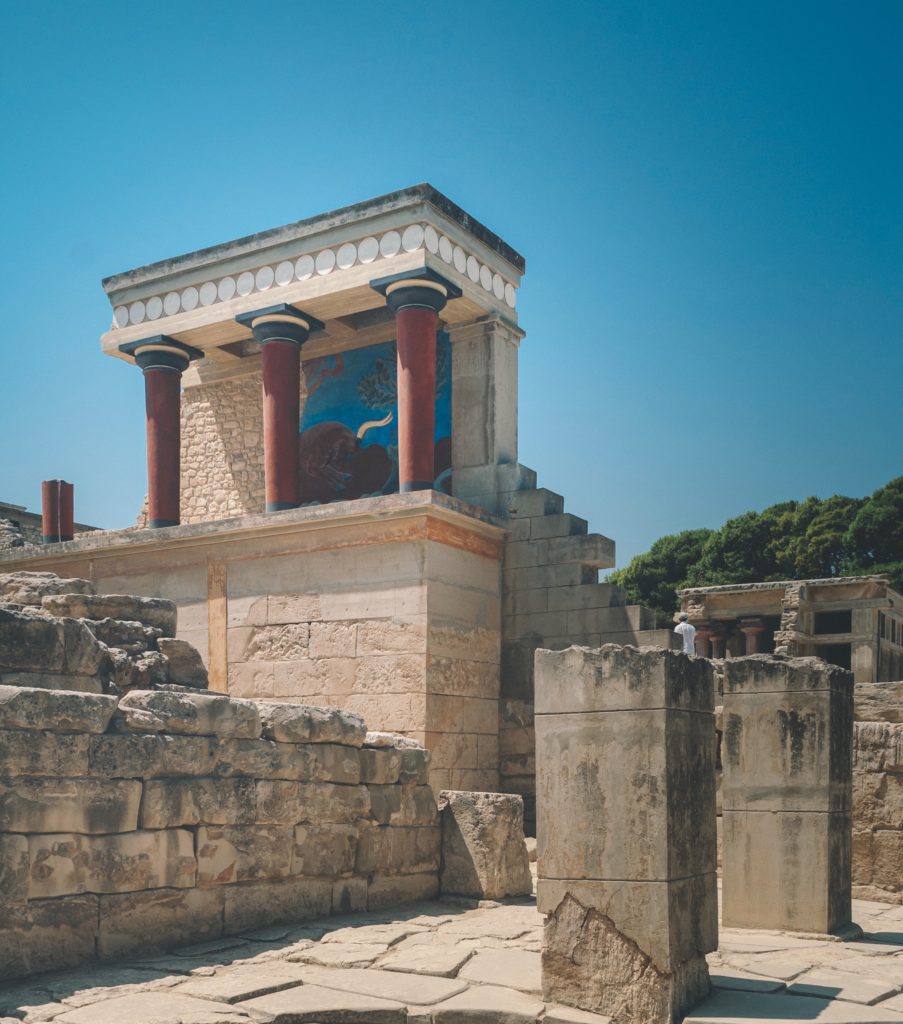 Against the blue sky stands the ruins of Knossos Palace with its red stone columns. 