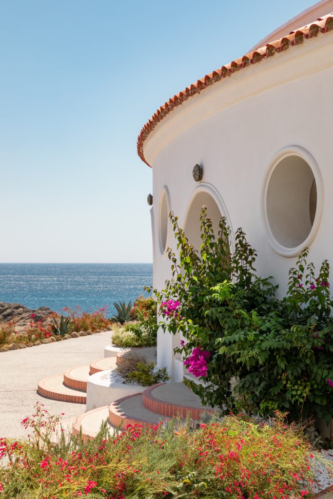 A white circular villa with a terracotta roof shrouded by pink flowers with the blue sea and sky in the background.