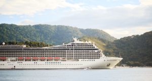 A large white cruise ship sails in front of tree covered hills.