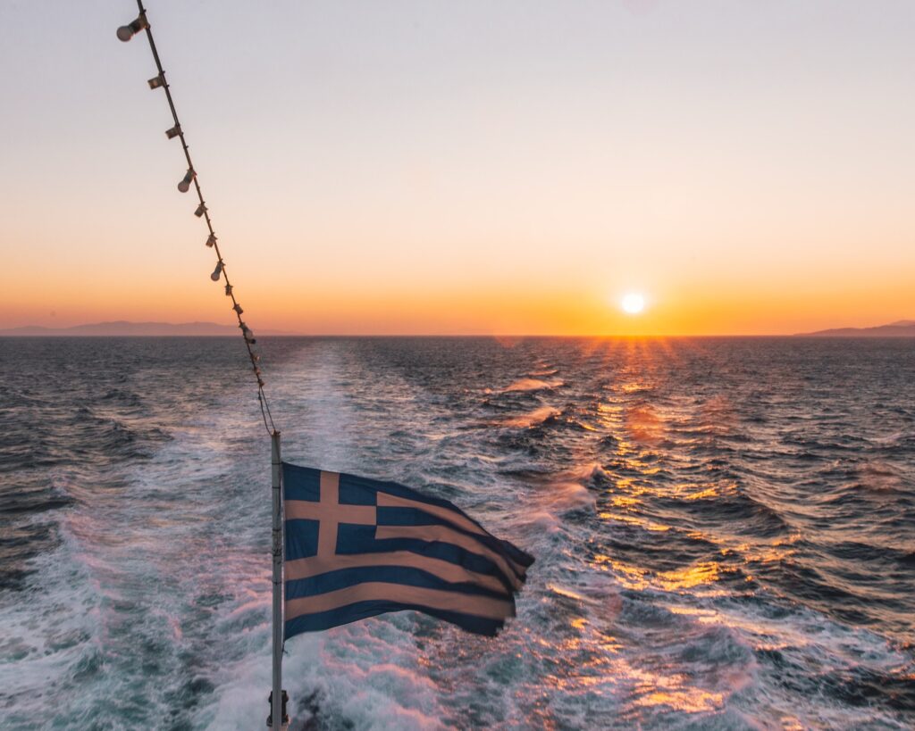 A greek flag flutters off the back of a ferry transfer to a greek island at sunset.