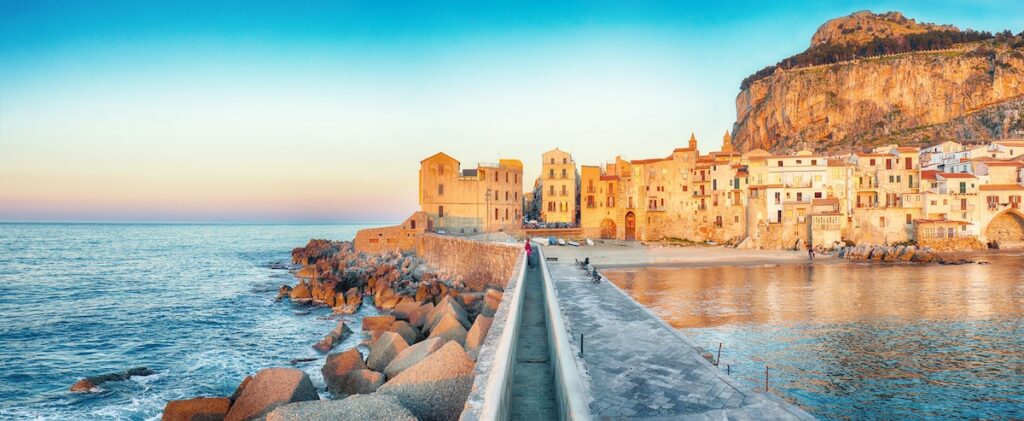 walled pathway over a sea of water with sand coloured buildings in the background of an old Sicilian town